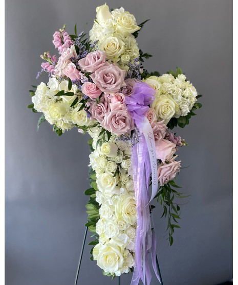 A cross-shaped sympathy arrangement of pink flowers with green accents, displayed on a standing easel, symbolizing faith, love, and remembrance.