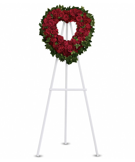 A heart-shaped wreath of all red flowers with green accents, displayed on a standing easel with an open center, symbolizing love, respect, and remembrance.