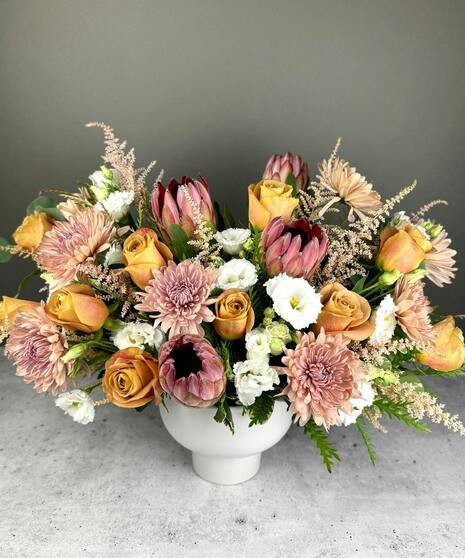 Boho arrangement with protea, roses, lisianthus, and textured blooms in blush, gold, ivory, and mauve tones.