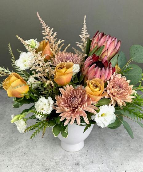 Boho arrangement with protea, roses, lisianthus, and textured blooms in blush, gold, ivory, and mauve tones.