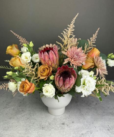 Boho arrangement with protea, roses, lisianthus, and textured blooms in blush, gold, ivory, and mauve tones.