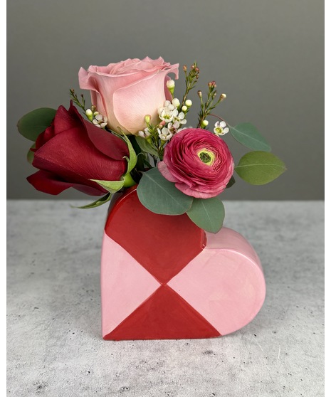 Small floral arrangement in a red and pink heart-shaped ceramic vase with romantic seasonal blooms.