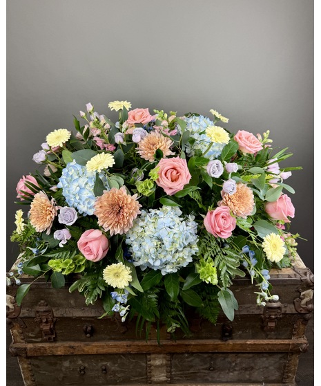 Pastel casket spray with seasonal flowers in soft pink, yellow, lavender, and green.