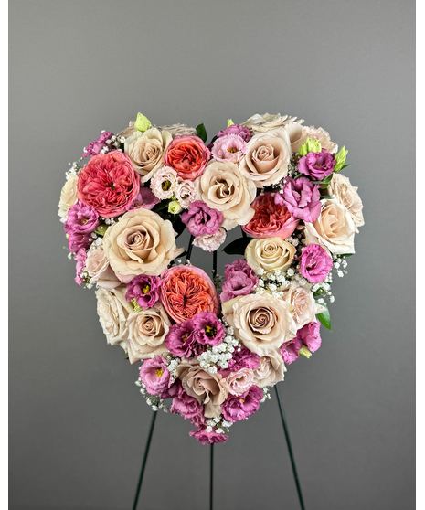 A heart-shaped wreath of blush and pink flowers with green accents, displayed on a standing easel with an open center, symbolizing love and grace