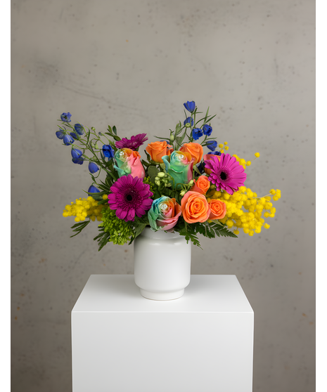 Bright colorful flower arrangement with rainbow roses and festive blooms in a white vase.