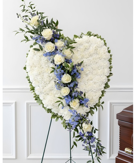 A heart-shaped sympathy wreath of blue and white flowers with green accents, displayed on a standing easel, symbolizing peace, love, and remembrance.