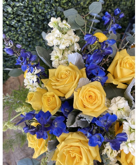 A heart-shaped wreath of blue, yellow, and white flowers with green accents, displayed on a standing easel with an open center, perfect for honoring Navy service.