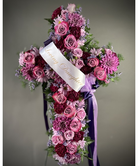A cross-shaped sympathy arrangement of pink and purple flowers with green accents, displayed on a standing easel, symbolizing faith, love, and remembrance.