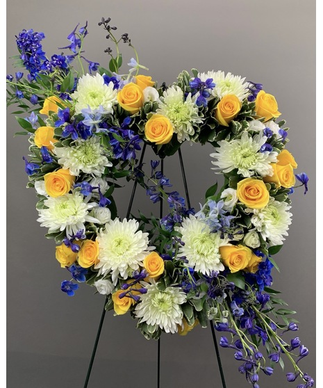 A heart-shaped wreath of blue, yellow, and white flowers with green accents, displayed on a standing easel with an open center, perfect for honoring Navy service.