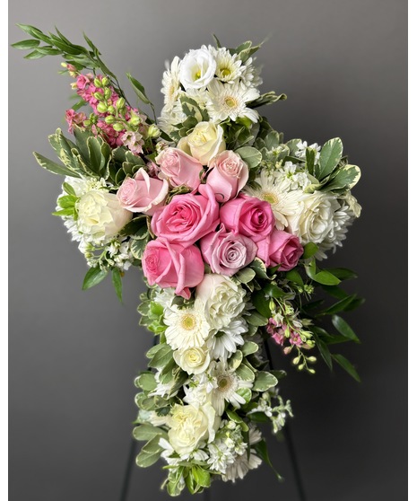 A cross-shaped sympathy arrangement of pink flowers with green accents, displayed on a standing easel, symbolizing faith, love, and remembrance.
