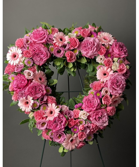 A heart-shaped wreath of blush and pink flowers with green accents, displayed on a standing easel with an open center, symbolizing love and grace