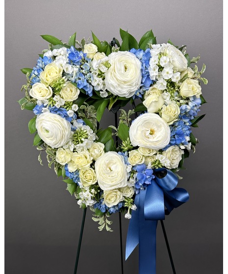 A heart-shaped wreath of white and blue flowers with green accents, displayed on a standing easel with an open center, symbolizing peace and love.