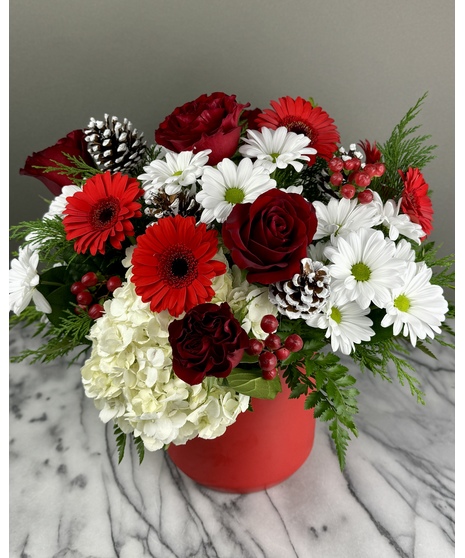 A festive holiday centerpiece featuring red roses, white daisies, red gerbera daisies, white hydrangeas, red berries, evergreen sprigs, and frosted pinecones in a red vase. 
