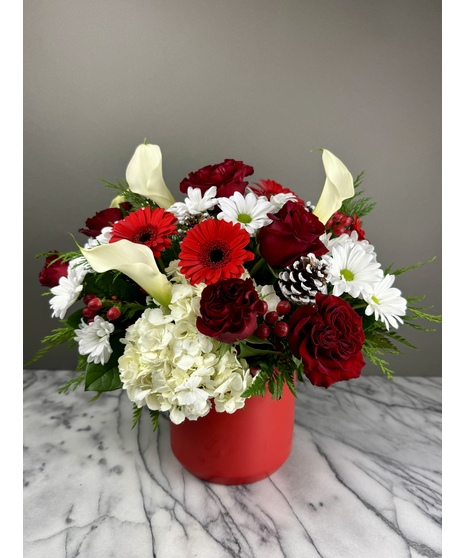 A festive holiday centerpiece featuring red roses, white daisies, red gerbera daisies, white hydrangeas, red berries, evergreen sprigs, and frosted pinecones in a red vase. 