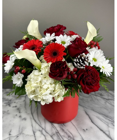 A festive holiday centerpiece featuring red roses, white daisies, red gerbera daisies, white hydrangeas, red berries, evergreen sprigs, and frosted pinecones in a red vase. 