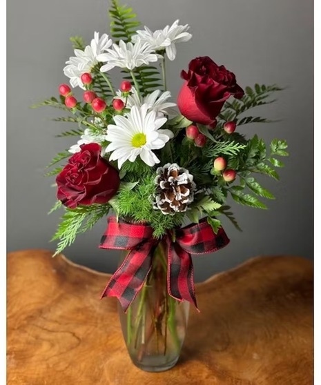 A holiday bouquet featuring red roses, white daisies, red berries, and evergreen sprigs, with a frosted pinecone and a red plaid bow, arranged in a clear glass vase.