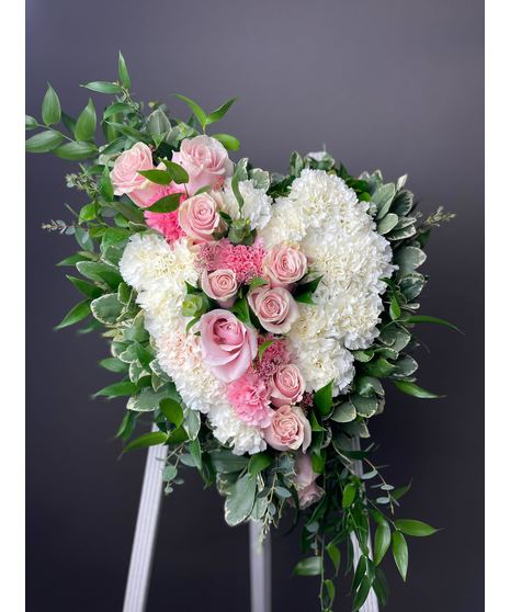 A heart-shaped sympathy wreath in shades of pink, blush, and white with green accents, displayed on a standing easel with a broken heart effect.