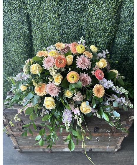 Pastel casket spray with peach, yellow, pink, and lavender blooms with lush greenery.