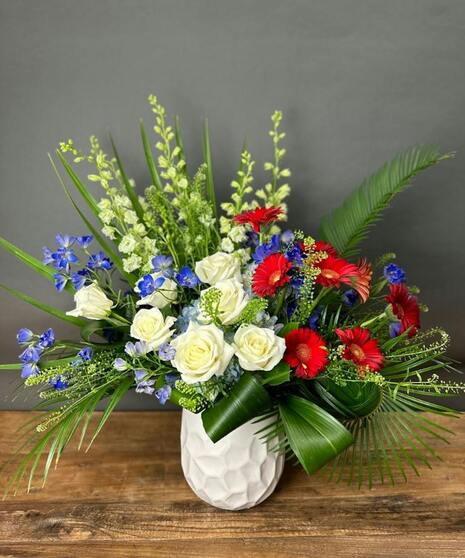 Red roses with white and blue blooms arranged in lush greenery for a patriotic sympathy tribute.