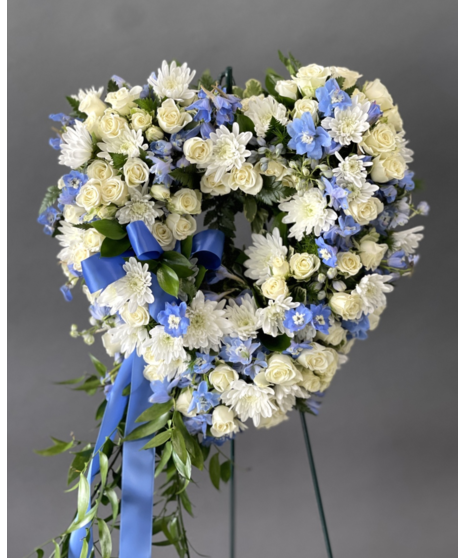 A heart-shaped wreath of white and blue flowers with green accents, displayed on a standing easel with an open center, symbolizing peace and love.