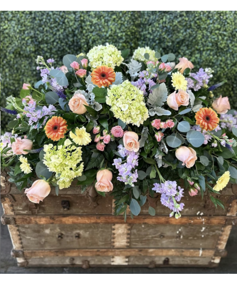 Pastel casket spray with seasonal flowers in soft pink, yellow, lavender, and green.