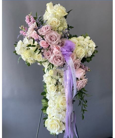 A cross-shaped sympathy arrangement of pink and purple flowers with green accents, displayed on a standing easel, symbolizing faith, love, and remembrance.