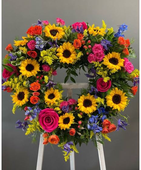 A heart-shaped wreath of vibrant, colorful seasonal flowers, displayed on a standing easel with an open center, perfect for celebrations or memorials.