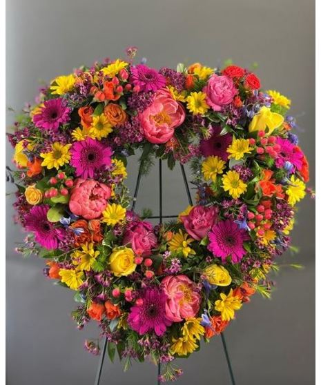 A heart-shaped wreath of vibrant, colorful seasonal flowers, displayed on a standing easel with an open center, perfect for celebrations or memorials.