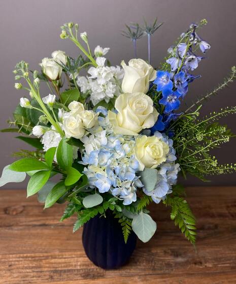 Blue and white floral arrangement with accent greenery in a glass or ceramic vase.