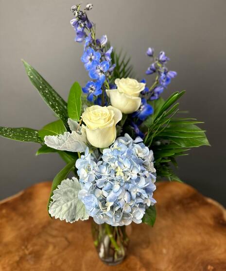Blue and white floral arrangement with accent greenery in a glass or ceramic vase.