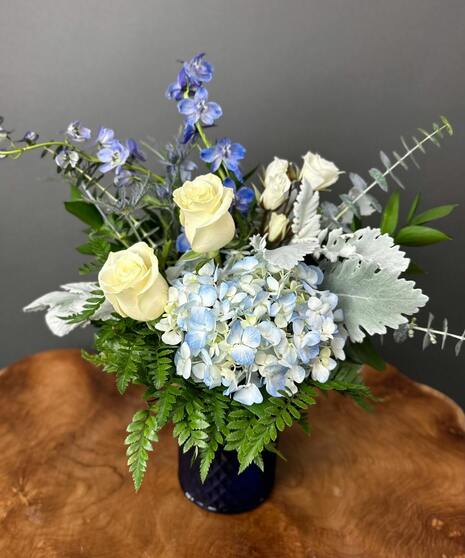 Blue and white floral arrangement with accent greenery in a glass or ceramic vase.