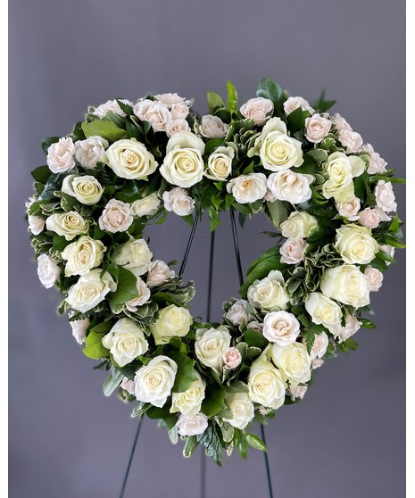 A heart-shaped wreath of pure white flowers, displayed on a standing easel with an open center, symbolizing peace, love, and remembrance.
