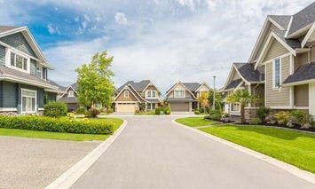 Suburban homes line tidy streets with well-manicured lawns and gardens, under a blue, partly cloudy sky, creating a quiet neighborhood scene.