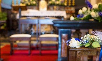 Flower arrangement rests on a wooden stand; background features two chairs on a red carpet in front of an ornate altar with white covering and abundant floral decorations nearby.