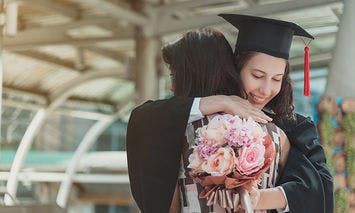A graduate in cap and gown embraces another person while holding a bouquet of flowers under an airy, roofed, outdoor structure.