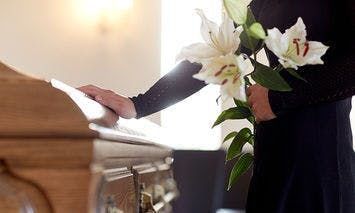 A person in black attire stands by a wooden casket, holding white lilies, in a softly-lit, indoor space, possibly a funeral home.
