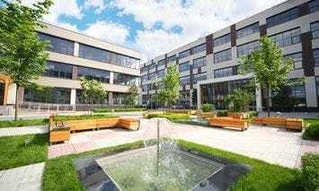 A modern courtyard features a central fountain surrounded by green grass, wooden benches, and young trees, with multi-story office buildings in the background under a partly cloudy sky.