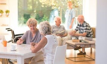 Elderly women converse at a table in a bright room, while a nurse attends to other elderly people sitting on a couch and chair in the background.