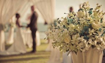 White and yellow flowers in the foreground with a blurred couple exchanging vows in a sunlit outdoor setting under a large white canopy in the background.