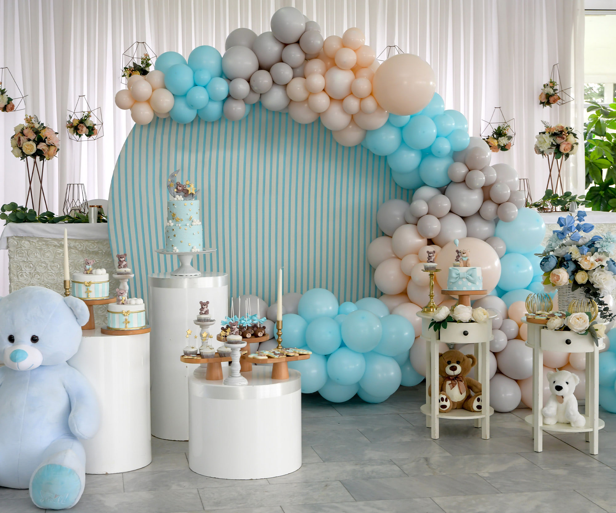 Baby shower dessert table with blue balloon arch, teddy bear decor, floral arrangements, and themed sweets display
